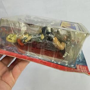 Peut inclure: Une figurine d'action emball&eacute;e repr&eacute;sentant un personnage &agrave; la peau jaune v&ecirc;tu de noir, sur une base rouge. La figurine est dans une coque en plastique transparent, avec un dos en carton rouge et bleu.