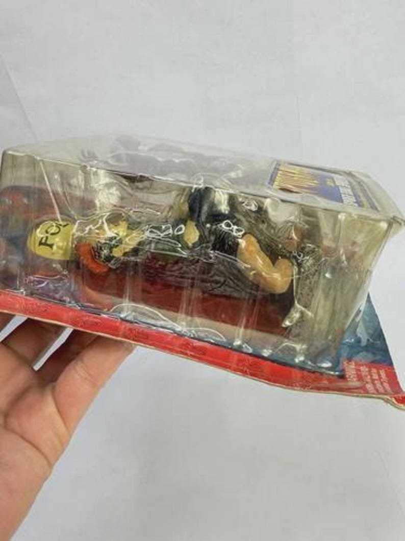Peut inclure: Une figurine d'action emball&eacute;e repr&eacute;sentant un personnage &agrave; la peau jaune v&ecirc;tu de noir, sur une base rouge. La figurine est dans une coque en plastique transparent, avec un dos en carton rouge et bleu.