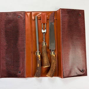 May include: A vintage carving set with a sharpening steel, fork, and knife, displayed in an open leather case. The case is brown with a textured, faux-snakeskin pattern. The knife handles are a light brown color.