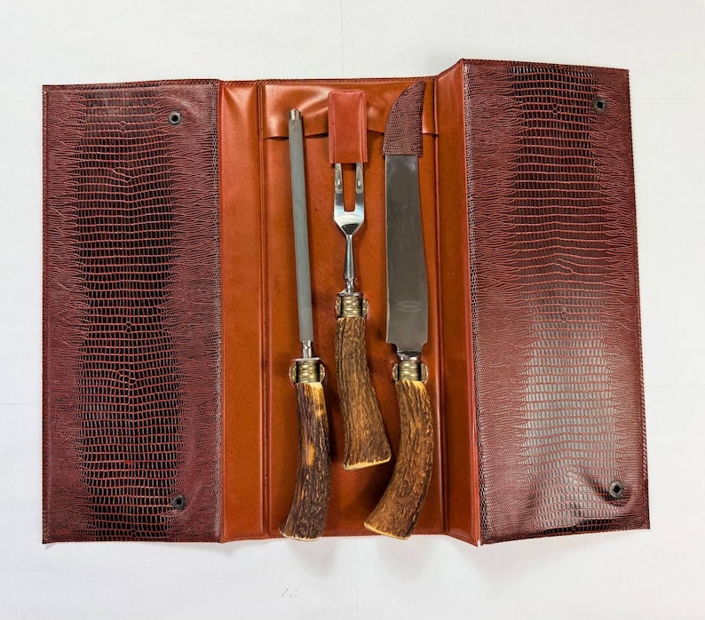 May include: A vintage carving set with a sharpening steel, fork, and knife, displayed in an open leather case. The case is brown with a textured, faux-snakeskin pattern. The knife handles are a light brown color.
