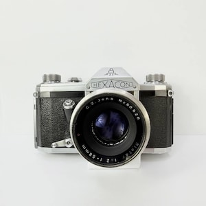 May include: A vintage Hexacon camera with a black leather body and silver accents. The lens is black with the text "C.Z. Jena" and "Nr. 60526" printed on it. The lens also has the text "f=58mm 1:2" printed on it.