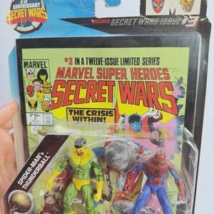 K&ouml;nnte beinhalten: Marvel Secret Wars Actionfiguren-Set mit Spider-Man und Thunderball. Die Verpackung enth&auml;lt den Text "25th Anniversary Secret Wars" und "Marvel Super Heroes Secret Wars". Das Comic-Cover-Artwork ist im Hintergrund sichtbar.
