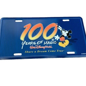 May include: A rectangular blue license plate featuring a vertical pinstripe pattern. The text reads 100 YEARS OF MAGIC Walt Disney World Share a Dream Come True. A graphic of Mickey Mouse is positioned to the right of the large orange 100.