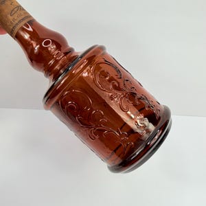 Puede incluir: Una botella de vidrio marr&oacute;n con un dise&ntilde;o floral en relieve. La botella tiene una base bulbosa y un cuello largo, con un tap&oacute;n de corcho. La botella es probablemente para fines decorativos.