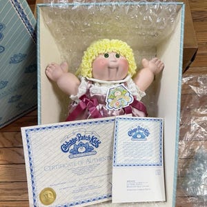May include: A Cabbage Patch Kids porcelain doll with curly yellow hair and green eyes, wearing a pink satin dress with white lace trim. The doll is inside a light blue box with bubble wrap and includes a Certificate of Authenticity document.