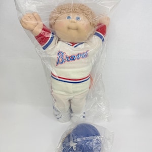 Puede incluir: Una muñeca Cabbage Patch Kids con cabello de lana marrón claro y ojos azules, que viste un uniforme de béisbol blanco con ribetes rojos y azules y la palabra Braves impresa en el pecho. La muñeca está sellada en una bolsa de plástico.