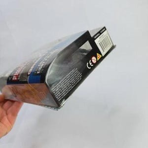 May include: A product box with a clear plastic front, partially visible. The box features a black and blue design with white text. The box is held at an angle, with a hand visible in the lower left corner.