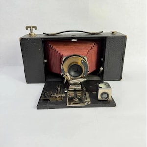 May include: A vintage black folding camera with a red bellows and a brass lens. The camera has a handle on top and a small viewfinder. The front panel is open, revealing the internal mechanisms.