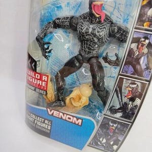 May include: Venom action figure in a clear plastic package. The figure is black with white spiderweb-like patterns and a long red tongue. The packaging includes the word "Venom" and images of the character.