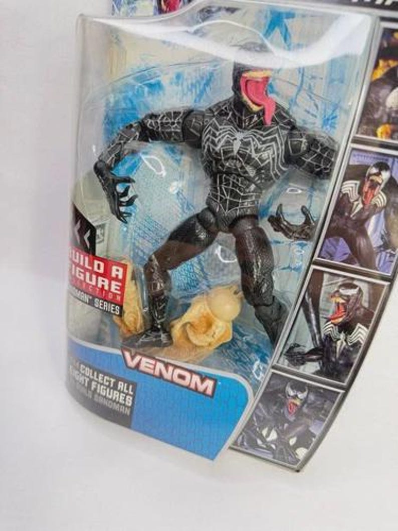 May include: Venom action figure in a clear plastic package. The figure is black with white spiderweb-like patterns and a long red tongue. The packaging includes the word "Venom" and images of the character.