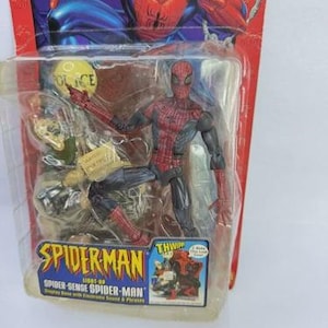 Peut inclure: Figurine articul&eacute;e de Spider-Man dans un emballage plastique. La figurine est rouge et noire, avec un costume d&eacute;taill&eacute;. L'emballage pr&eacute;sente le logo Spider-Man et le texte "Spider-Sense Spider-Man". L'arri&egrave;re-plan montre une illustration de Spider-Man.
