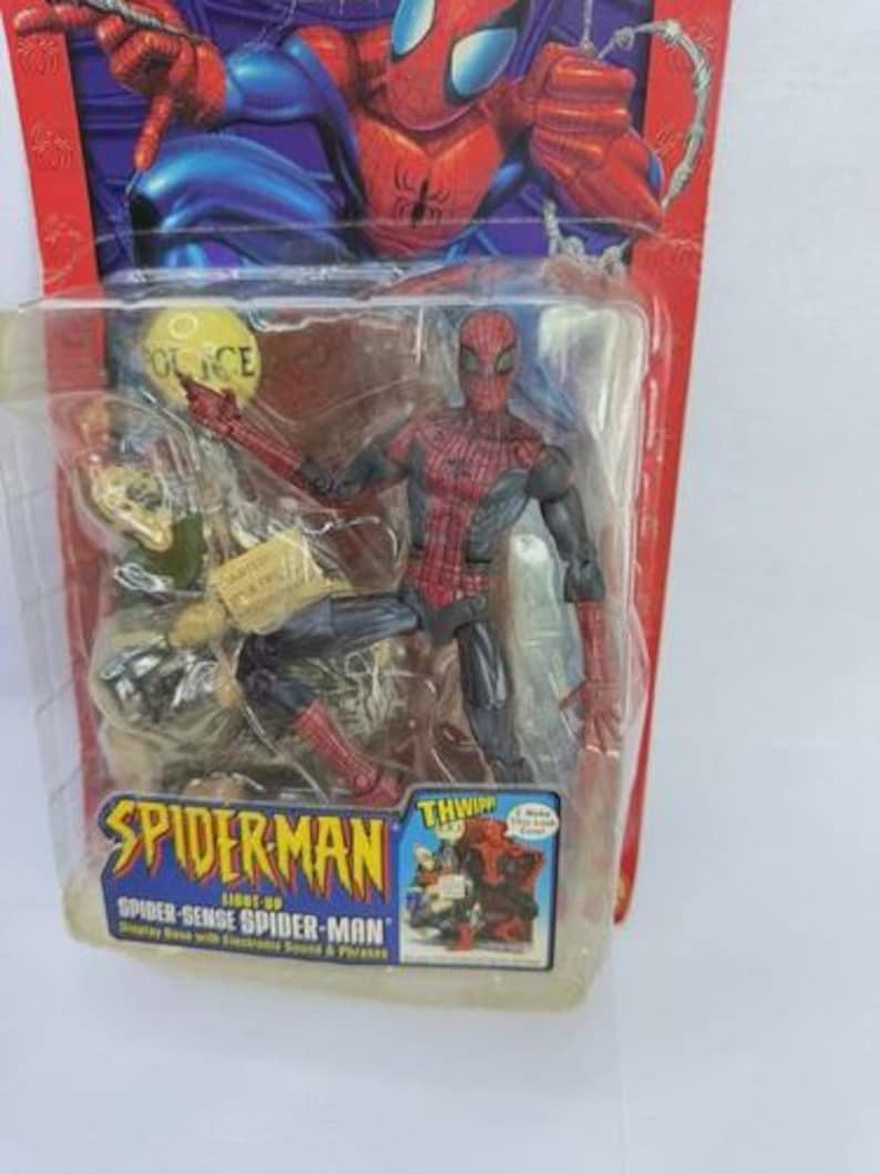 Peut inclure: Figurine articul&eacute;e de Spider-Man dans un emballage plastique. La figurine est rouge et noire, avec un costume d&eacute;taill&eacute;. L'emballage pr&eacute;sente le logo Spider-Man et le texte "Spider-Sense Spider-Man". L'arri&egrave;re-plan montre une illustration de Spider-Man.