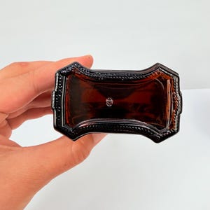 Puede incluir: Una botella de vidrio de color &aacute;mbar oscuro con un dise&ntilde;o rectangular decorativo. La botella tiene un borde detallado y adornado y una forma ligeramente curvada. La botella est&aacute; sostenida por una mano sobre un fondo blanco.