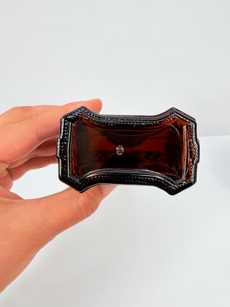 Puede incluir: Una botella de vidrio de color &aacute;mbar oscuro con un dise&ntilde;o rectangular decorativo. La botella tiene un borde detallado y adornado y una forma ligeramente curvada. La botella est&aacute; sostenida por una mano sobre un fondo blanco.
