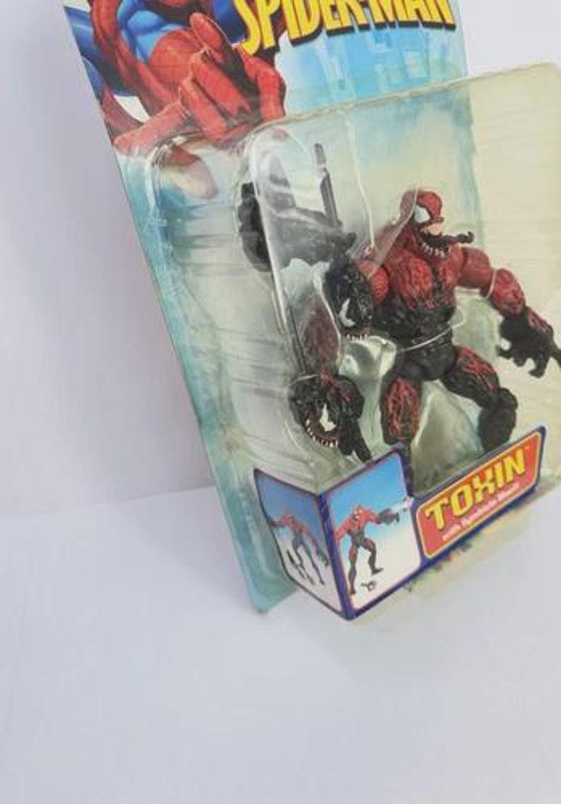 Puede incluir: Figura de acci&oacute;n de Toxin roja y negra de la serie Spider-Man, empaquetada en un bl&iacute;ster transparente. El embalaje presenta gr&aacute;ficos de Spider-Man y la palabra "TOXIN" en amarillo y rojo.