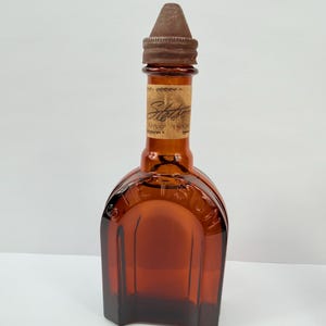 Puede incluir: Botella de vidrio color &aacute;mbar con tap&oacute;n marr&oacute;n. La botella tiene una base rectangular y una parte superior redondeada con la palabra "Stetson" visible. Una etiqueta est&aacute; adherida al cuello de la botella.