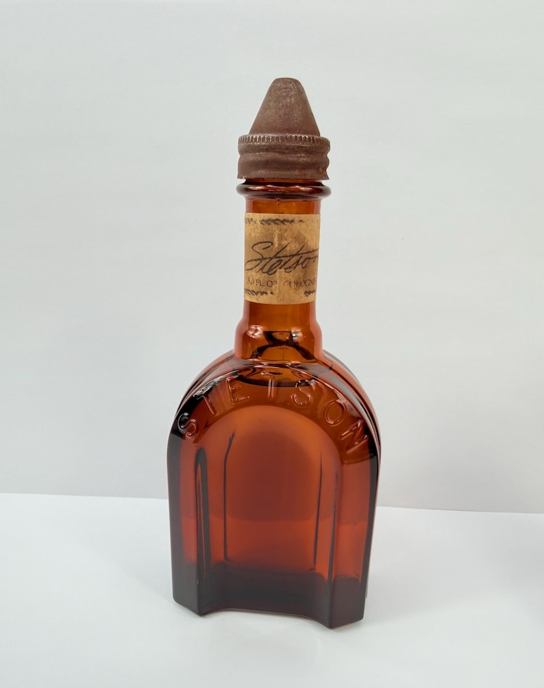 Puede incluir: Botella de vidrio color &aacute;mbar con tap&oacute;n marr&oacute;n. La botella tiene una base rectangular y una parte superior redondeada con la palabra "Stetson" visible. Una etiqueta est&aacute; adherida al cuello de la botella.