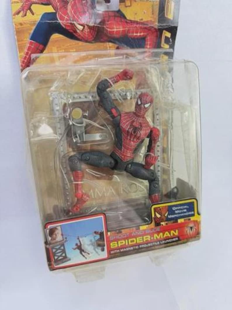 May include: Spider-Man action figure in a clear plastic package. The figure is red and black, in a dynamic pose. The packaging includes the text "SHOOT AND SLIDE SPIDER-MAN" and "Official Movie Merchandise".