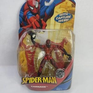 May include: A packaged Carnage action figure from the Spider-Man series. The figure is red and black, with a detailed design. The packaging features Spider-Man artwork and the text "WITH CAPTURE WEBS!" and "SUPER VILLAIN". The packaging is blue and yellow.