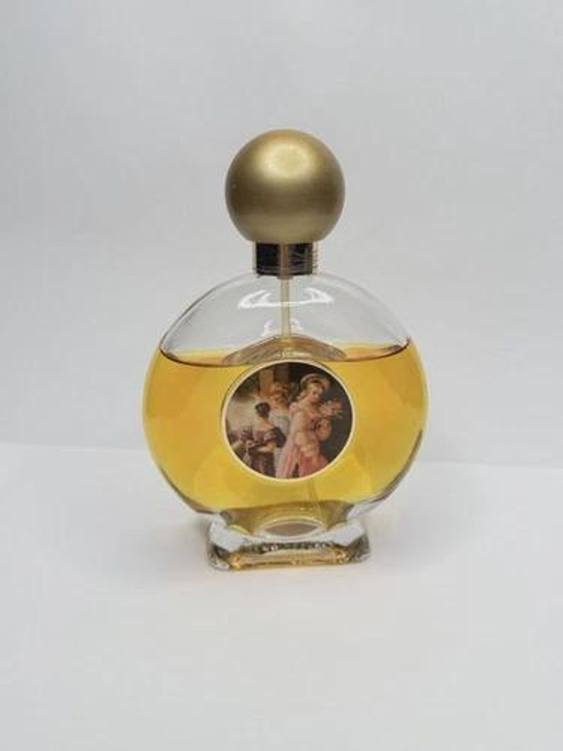 May include: A vintage perfume bottle with a clear, rounded glass body and a gold-colored spherical stopper. The bottle contains a golden liquid and features a circular label with a classical painting. The base is a clear, rectangular shape.