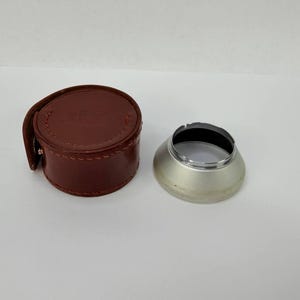 Puede incluir: Un estuche de cuero marr&oacute;n para lentes de c&aacute;mara y un parasol plateado. El estuche es cil&iacute;ndrico con cierre de bot&oacute;n a presi&oacute;n. El parasol es met&aacute;lico con interior negro y forma ancha y acampanada. Equipo de fotograf&iacute;a.