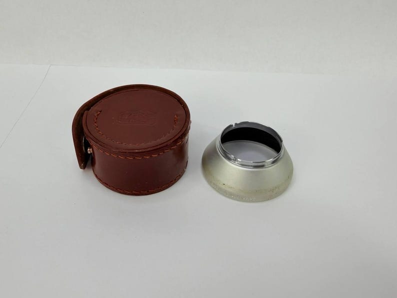 Puede incluir: Un estuche de cuero marr&oacute;n para lentes de c&aacute;mara y un parasol plateado. El estuche es cil&iacute;ndrico con cierre de bot&oacute;n a presi&oacute;n. El parasol es met&aacute;lico con interior negro y forma ancha y acampanada. Equipo de fotograf&iacute;a.