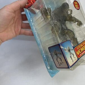 May include: A Stealth Venom action figure in a clear plastic package. The packaging features Spider-Man graphics and the Marvel logo. A red "Try Me!" sticker is visible on the package.