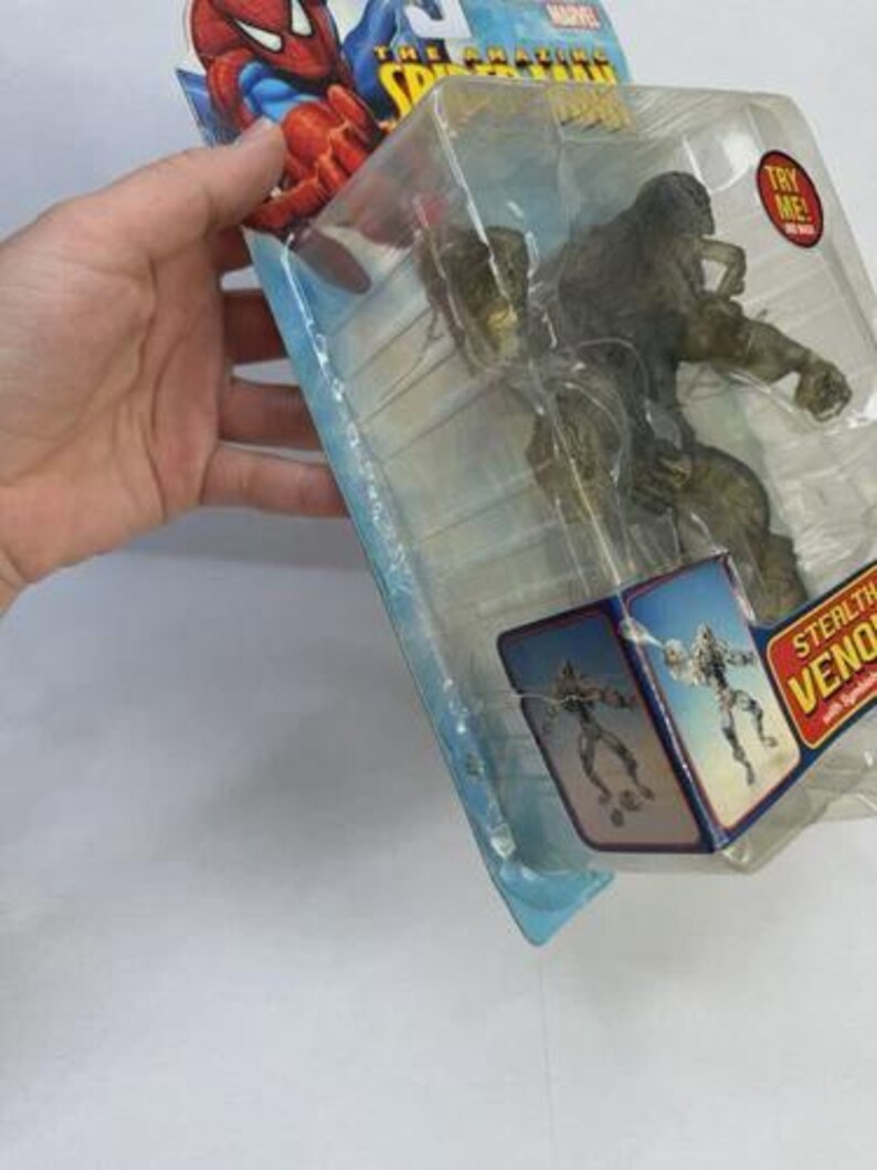 May include: A Stealth Venom action figure in a clear plastic package. The packaging features Spider-Man graphics and the Marvel logo. A red "Try Me!" sticker is visible on the package.