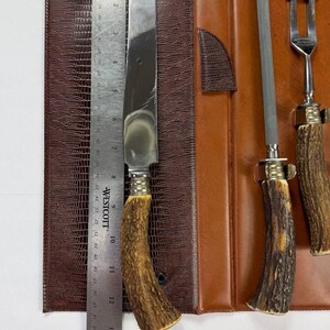 May include: A vintage steak knife set with a brown leather case. The set includes a large knife, a sharpening steel, and a fork. The knife blade measures approximately 10 inches. The handles are made of a brown, textured material.