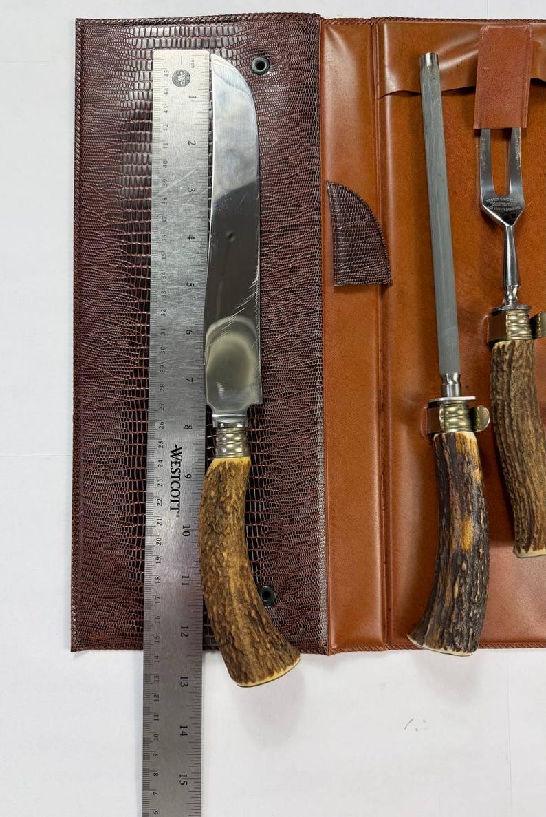May include: A vintage steak knife set with a brown leather case. The set includes a large knife, a sharpening steel, and a fork. The knife blade measures approximately 10 inches. The handles are made of a brown, textured material.
