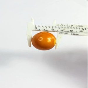 以下が含まれることがあります： 琥珀色の楕円形のビーズが白いノギスで測定されています。ビーズは光沢のある仕上げで、半透明の素材でできているようです。ノギスはセンチメートル単位の測定値を表示します。