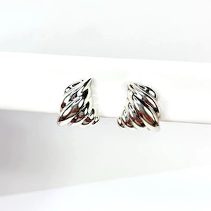 Puede incluir: Un par de pendientes de aro plateados. Cada pendiente presenta un diseño retorcido en forma de cuerda, creando una superficie texturizada y reflectante. Los pendientes están colocados sobre un fondo blanco brillante. Joyería.