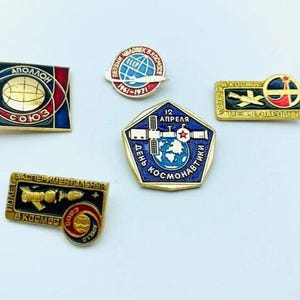May include: A collection of vintage space-themed pins. The pins feature various designs, including a globe, rockets, and the words "Apollo" and "Soyuz." The pins are in various shapes and colors, with gold, red, blue, and black accents.