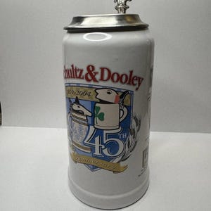 Peut inclure: Chope à bière en céramique blanche avec un couvercle et une poignée argentés. La chope porte l'inscription "Schultz & Dooley" en rouge et bleu, ainsi qu'un graphisme célébrant un 45e anniversaire. Le design comprend des personnages de dessins animés et des épis de blé.