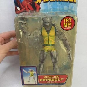 May include: Sealed action figure of Howling Manwolf from The Amazing Spider-Man series. The figure is gray and yellow, with articulated limbs and a wolf-like head. The packaging includes the text "TRY ME!" and "HOWLING MANWOLF".