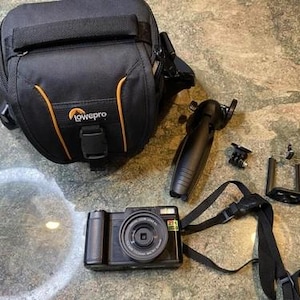 Op de afbeelding: Een zwarte camera met lens en riem, een zwarte cameratas met oranje accenten, een klein statief en een telefoonhouder zijn op een oppervlak geplaatst. De cameratas heeft het logo "lowepro".