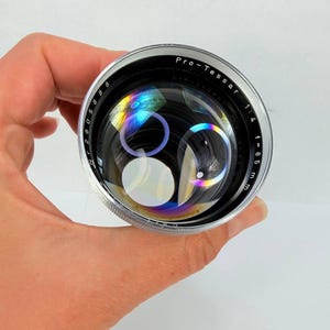Puede incluir: Un objetivo de c&aacute;mara Pro-Tessar plateado y negro con el texto "Pro-Tessar 1:4 f=85 mm". La lente tiene una superficie reflectante con refracciones de luz de color arco&iacute;ris. La lente se sostiene en una mano.