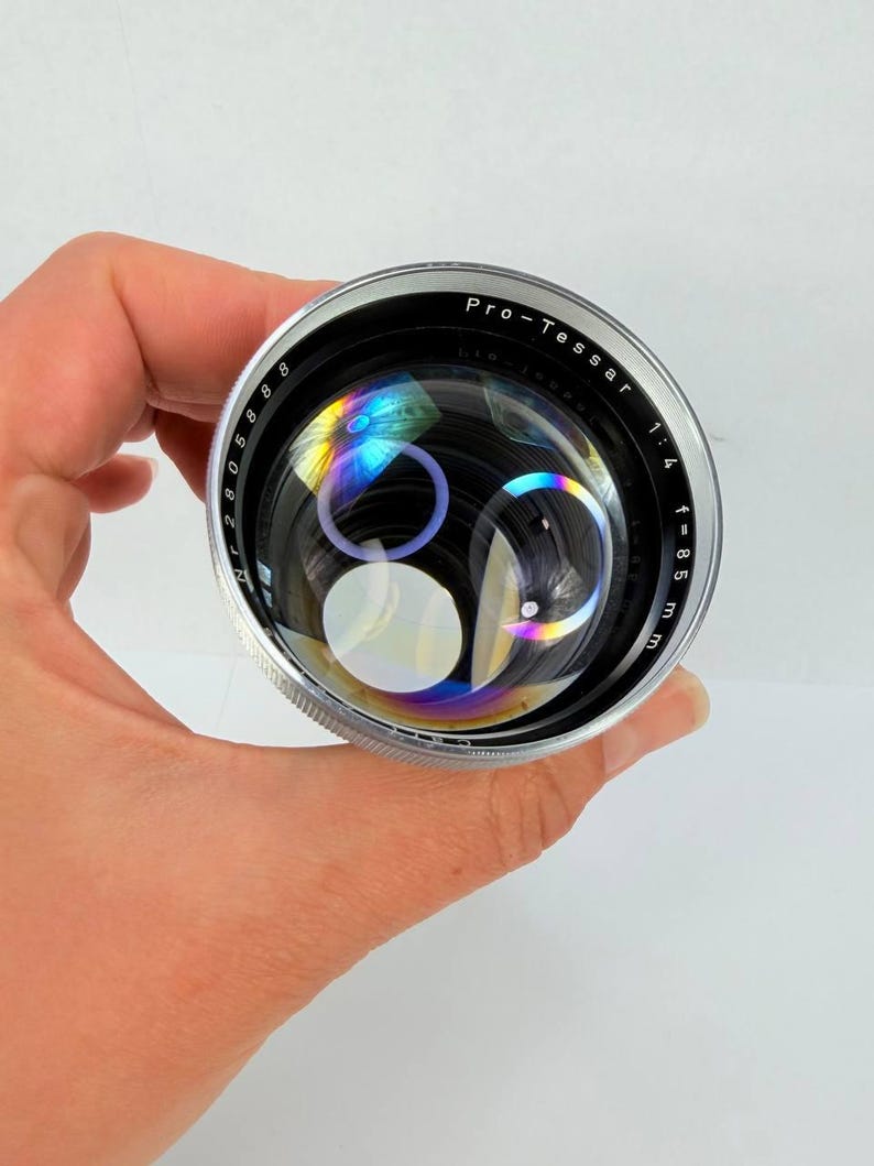 Puede incluir: Un objetivo de c&aacute;mara Pro-Tessar plateado y negro con el texto "Pro-Tessar 1:4 f=85 mm". La lente tiene una superficie reflectante con refracciones de luz de color arco&iacute;ris. La lente se sostiene en una mano.