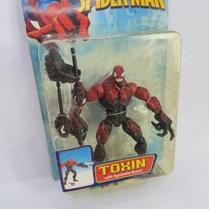 Puede incluir: Figura de acci&oacute;n de Toxin de la serie The Amazing Spider-Man, empaquetada en una funda de pl&aacute;stico transparente. La figura es roja y negra, con un dise&ntilde;o detallado. El embalaje presenta ilustraciones de Spider-Man y el logotipo de Marvel.