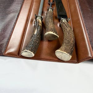 May include: A brown leather case with a reptile-like texture holds three steak knives. The knives have antler handles and silver-colored bolsters. The case is open, revealing the knives and a brown leather interior.