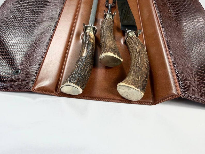 May include: A brown leather case with a reptile-like texture holds three steak knives. The knives have antler handles and silver-colored bolsters. The case is open, revealing the knives and a brown leather interior.