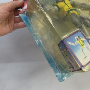 May include: A packaged action figure of a gray-skinned character in a yellow and green outfit. The figure is in a clear plastic package with a blue and red card featuring a character illustration. The packaging is held by a hand.