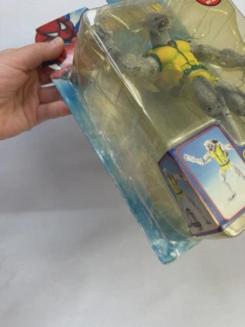 May include: A packaged action figure of a gray-skinned character in a yellow and green outfit. The figure is in a clear plastic package with a blue and red card featuring a character illustration. The packaging is held by a hand.