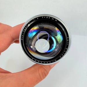 Puede incluir: Un objetivo de c&aacute;mara Carl Zeiss Pro-Tessar plateado y negro con una distancia focal de 85 mm y una apertura de f/1.4. La lente tiene una superficie reflectante con refracciones de luz de colores arco&iacute;ris.