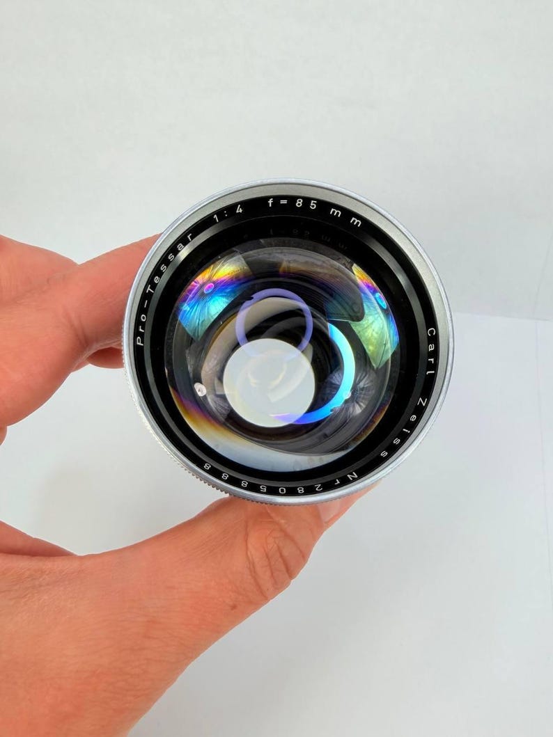 Puede incluir: Un objetivo de c&aacute;mara Carl Zeiss Pro-Tessar plateado y negro con una distancia focal de 85 mm y una apertura de f/1.4. La lente tiene una superficie reflectante con refracciones de luz de colores arco&iacute;ris.