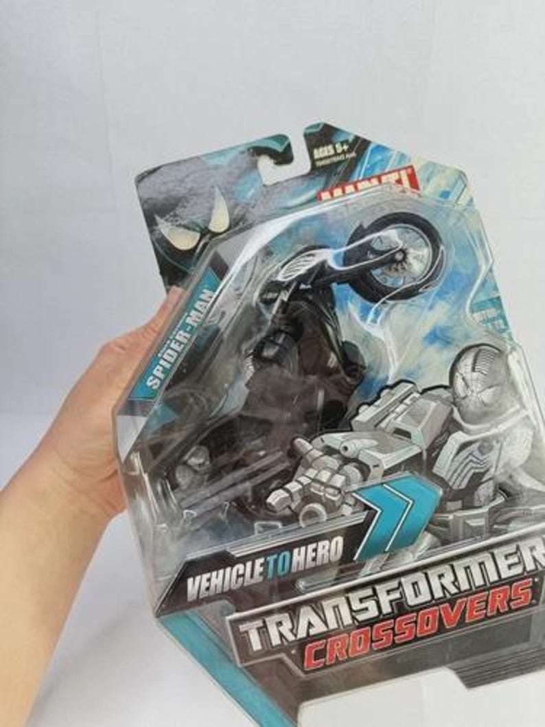 Puede incluir: Un juguete de Spider-Man Transformer Crossovers en su embalaje original. El juguete es una motocicleta negra con una figura de Spider-Man. El embalaje incluye el texto "Vehicle to Hero" y "Transformers Crossovers". El juguete es para mayores de 5 a&ntilde;os.