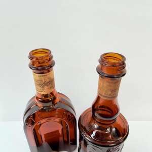 Puede incluir: Dos botellas de vidrio color &aacute;mbar con corchos. Una botella tiene una base cuadrada y la otra una base redonda con dise&ntilde;os florales en relieve. Ambas tienen etiquetas de papel con texto descolorido. Un tap&oacute;n de botella blanco est&aacute; en primer plano.