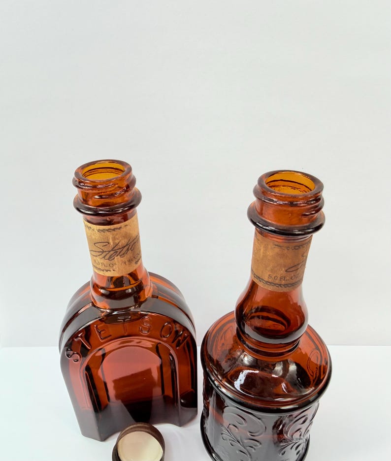 Puede incluir: Dos botellas de vidrio color &aacute;mbar con corchos. Una botella tiene una base cuadrada y la otra una base redonda con dise&ntilde;os florales en relieve. Ambas tienen etiquetas de papel con texto descolorido. Un tap&oacute;n de botella blanco est&aacute; en primer plano.