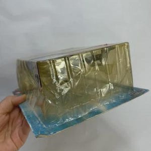 May include: A rectangular, sealed package in clear plastic. The package is held by a hand. The packaging has a blue and white border. The contents of the package are not visible.