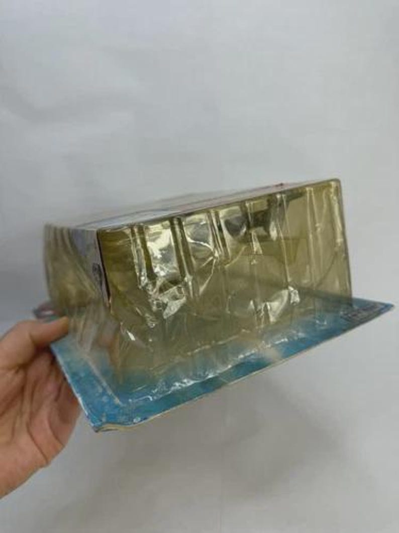 May include: A rectangular, sealed package in clear plastic. The package is held by a hand. The packaging has a blue and white border. The contents of the package are not visible.